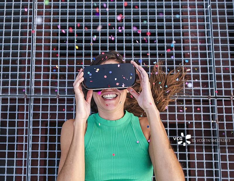 快乐的女人戴着虚拟现实眼镜躺在金属人行道上看五彩纸屑图片素材