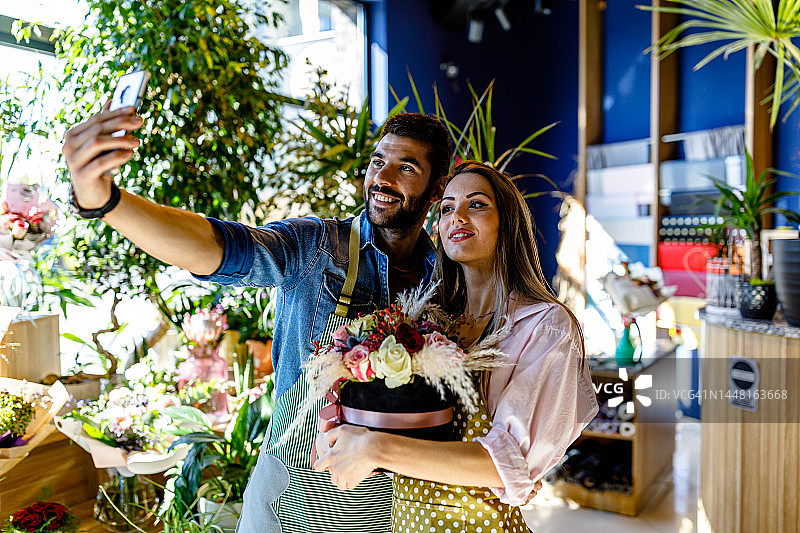 一对年轻漂亮的花匠夫妇在花店里用手机自拍。图片素材