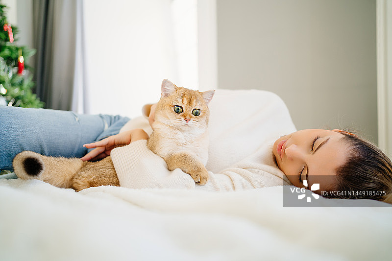 纯种猫的行为。懒散和放松的金发女人与马尾辫拥抱和抚摸她的姜猫，躺在白色的床上图片素材