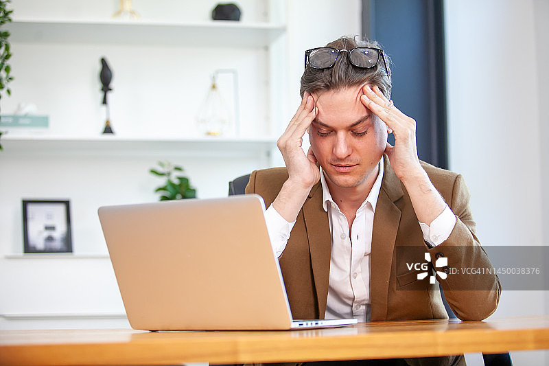 疲惫的商人头痛在办公室，感觉生病在工作与笔记本电脑和书在桌子上。图片素材