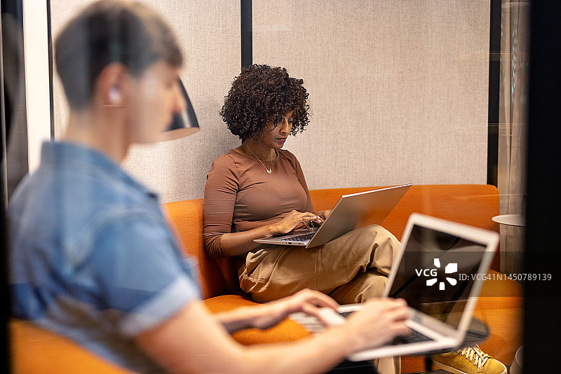 一名非洲妇女和一名同事坐在小隔间里用笔记本电脑工作图片素材