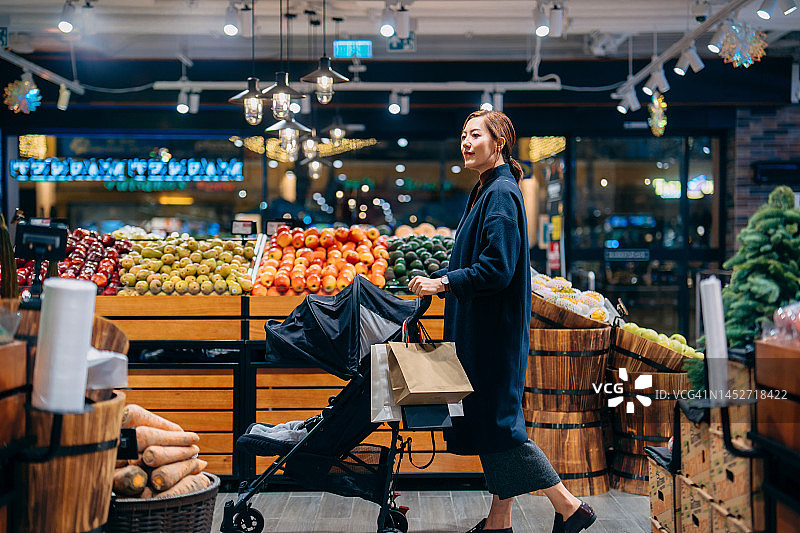 年轻的亚洲母亲推着婴儿车，带着她的孩子在超市购物。沿着水果和蔬菜走道走。日常购物。健康饮食方式图片素材