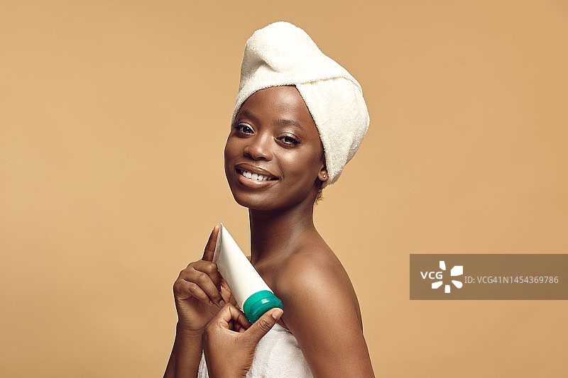 美丽的非洲裔妇女有干净的皮肤穿在身上的毛巾和水疗头巾持有保湿管。图片素材