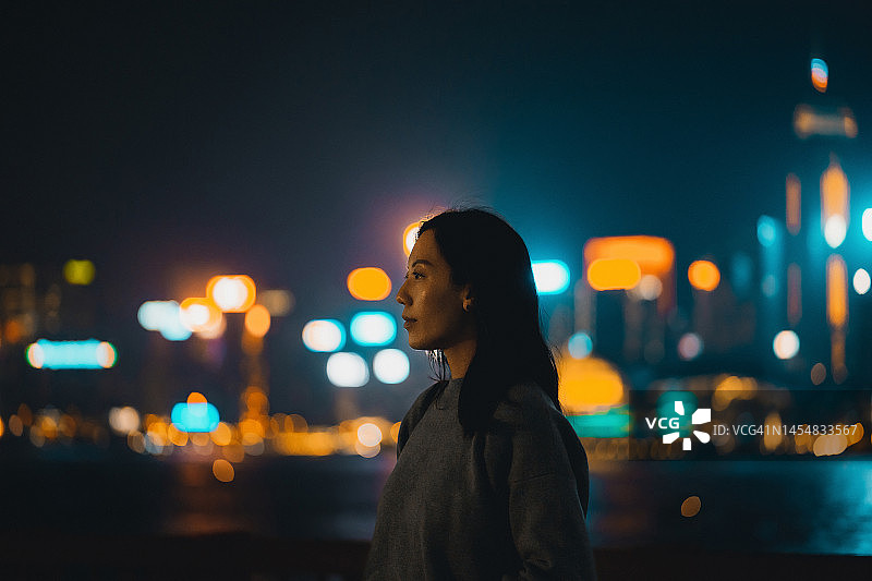一位年轻的亚洲女子站在维多利亚港的海滨长廊旁看向别处，夜晚的背景是明亮多彩的城市景观散景灯图片素材