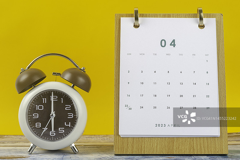 日历台2023:4月是组织者计划和截止日期的月份，桌子上有一个闹钟，背景是黄色的纸。图片素材