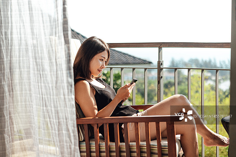 年轻的亚洲女子在阳台上使用智能手机放松图片素材