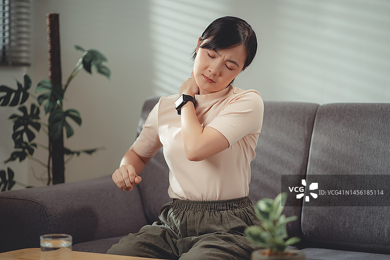 亚洲女性坐在家里客厅的沙发上脖子痛。图片素材
