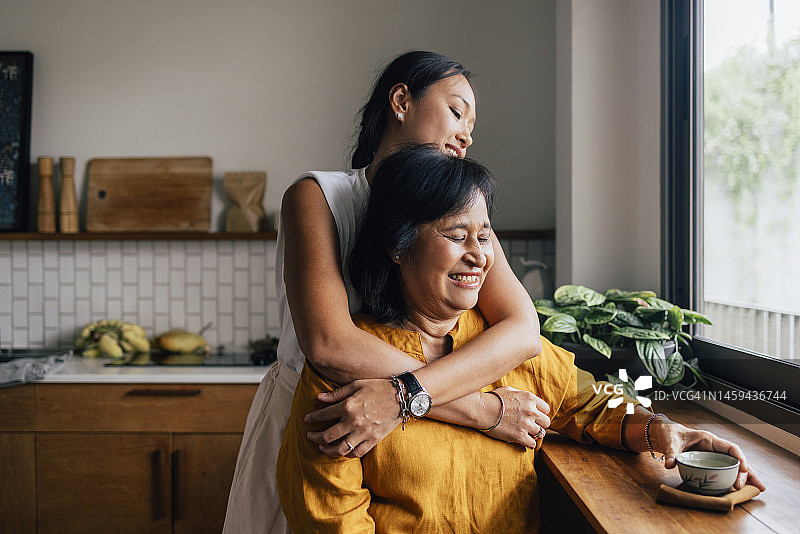 一个快乐美丽的女人拥抱着坐在厨房喝茶的妈妈图片素材