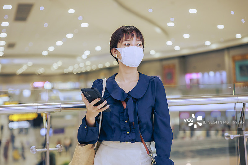 年轻的亚洲女商人戴着口罩站在机场离境区用手机看电子邮件。商务旅行的概念。图片素材