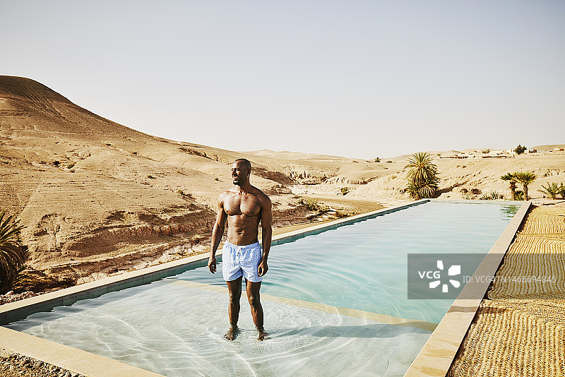宽镜头微笑的男人站在游泳池，而放松在沙漠营地图片素材