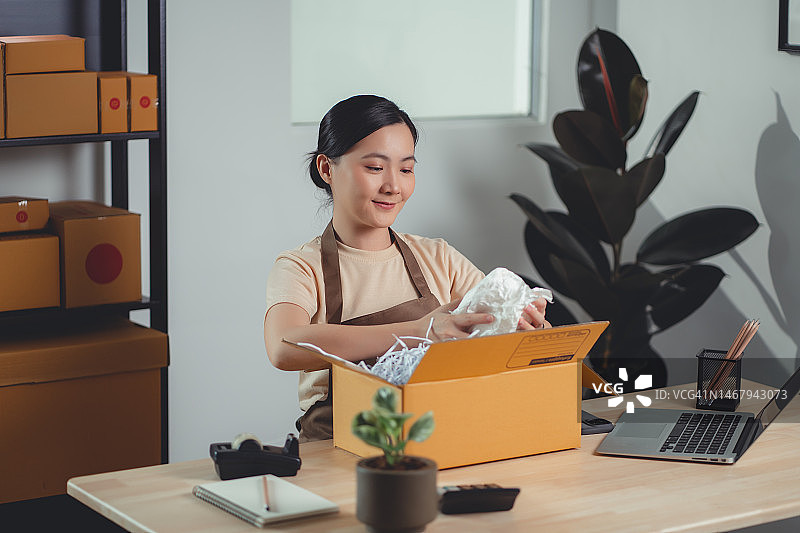 亚洲女网上卖家开心地微笑着在家里办公室包装产品。图片素材