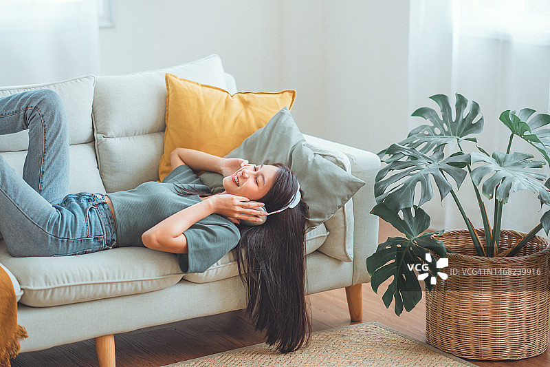 快乐的亚洲女人坐在家里的沙发上听手机里的音乐，微笑的女孩在早上戴着耳机放松，放松的时间。图片素材