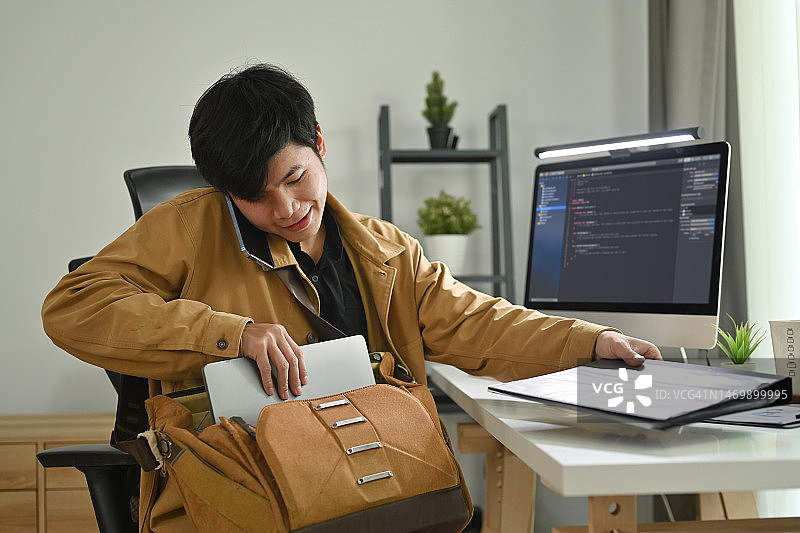 微笑的亚洲男性程序员在工作场所打电话，把他的东西公文包装进包里。图片素材