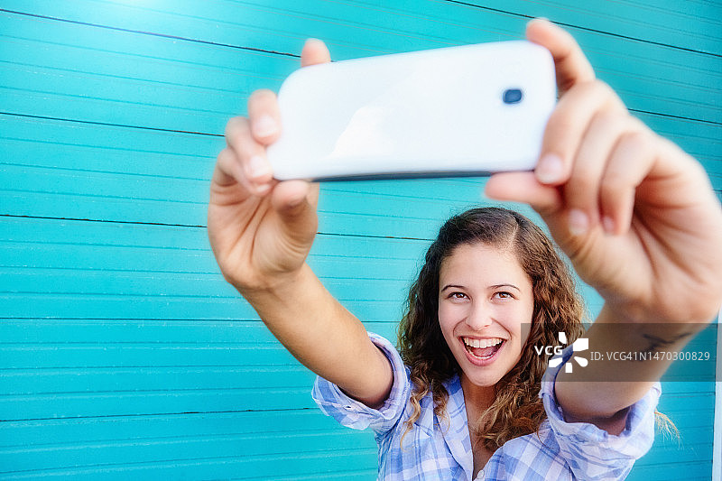 一名兴奋的年轻女子在户外靠在一扇绿松石色的卷帘门上，张开双臂，拿着手机自拍图片素材