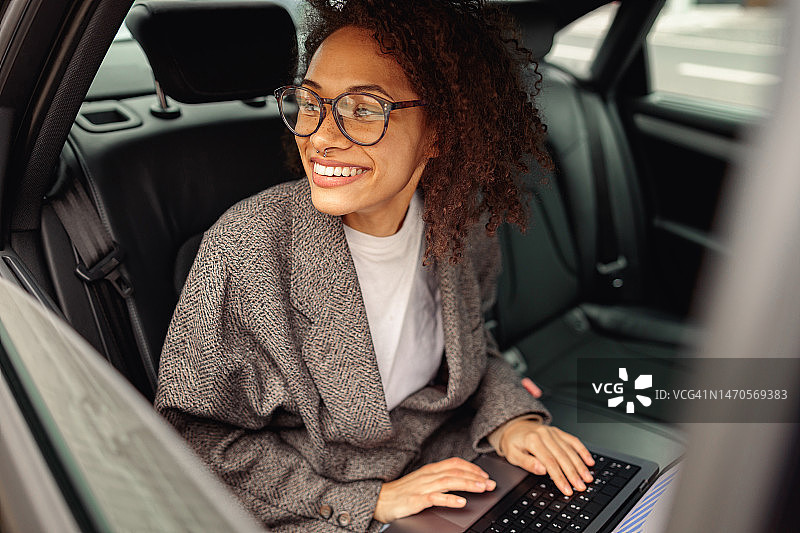 微笑的女程序员在笔记本电脑上工作，坐在汽车后座上班，看着窗户图片素材