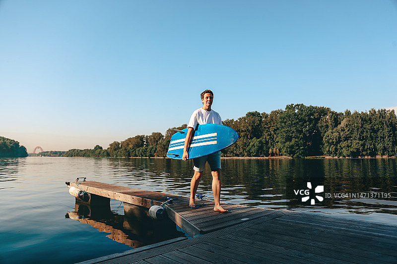 中年成年女性冲浪者走在木制码头与她的蓝色冲浪板在阳光明媚的夏日早晨图片素材