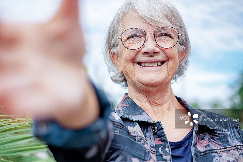 快乐迷人的老年女性在公园里散步，用手机自拍。戴着眼镜，满头白发，面带微笑的老太太拿着手机在户外享受自由图片素材