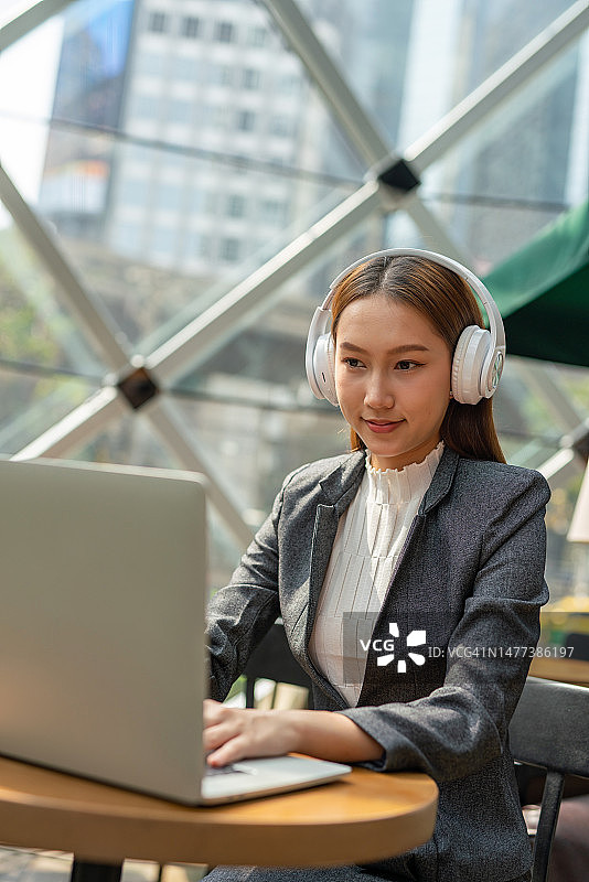 混合式工作风格。年轻的亚洲女性成年人与笔记本电脑在咖啡馆工作。戴白色耳机的女商人图片素材
