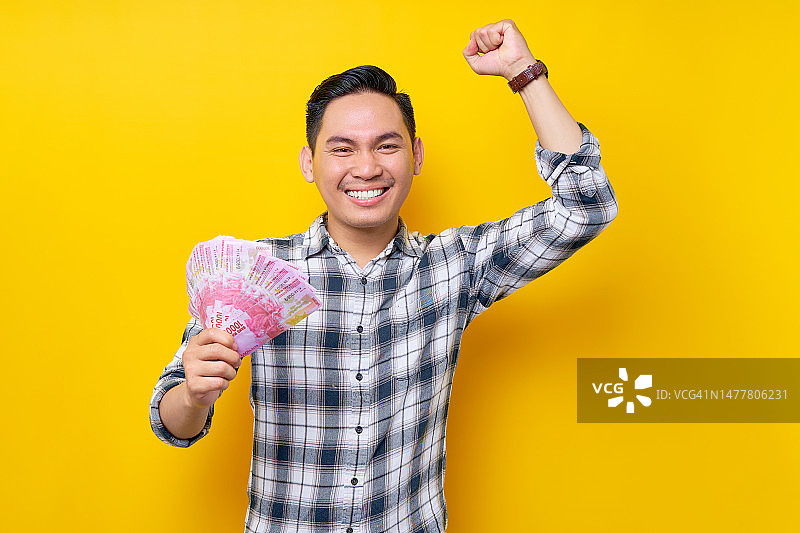 兴奋的年轻亚洲男子的肖像，穿着格子衬衫，拿着印尼盾钞票，并在黄色背景上展示胜利者的姿态。人们的生活方式理念图片素材