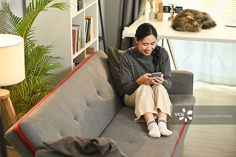满意的亚洲女子在网上聊天，在客厅放松时在手机上阅读信息。图片素材