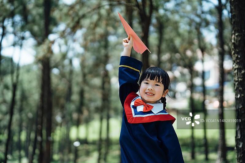 一个微笑的亚洲女孩的肖像，穿着毕业礼服，拿着纸飞机，看着相机，在一个阳光明媚的日子里，站在大自然中，背景是树木。保持自由和创造性。幼儿园。教育、学生和毕业观念图片素材