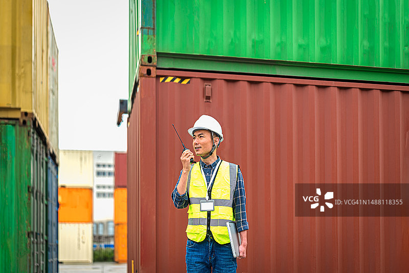 一名亚洲男性管理和运输控制工程师站在集装箱前，通过对讲机相互沟通。图片素材