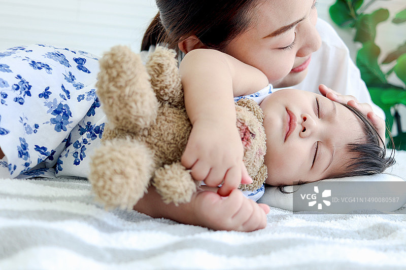 可爱的蹒跚学步的女婴女婴睡觉时抱着床上的泰迪熊玩具，年轻漂亮的妈妈温柔地抚摸熟睡的女儿孩子，妈妈安慰熟睡的孩子父母在家照顾孩子，爱护家庭图片素材