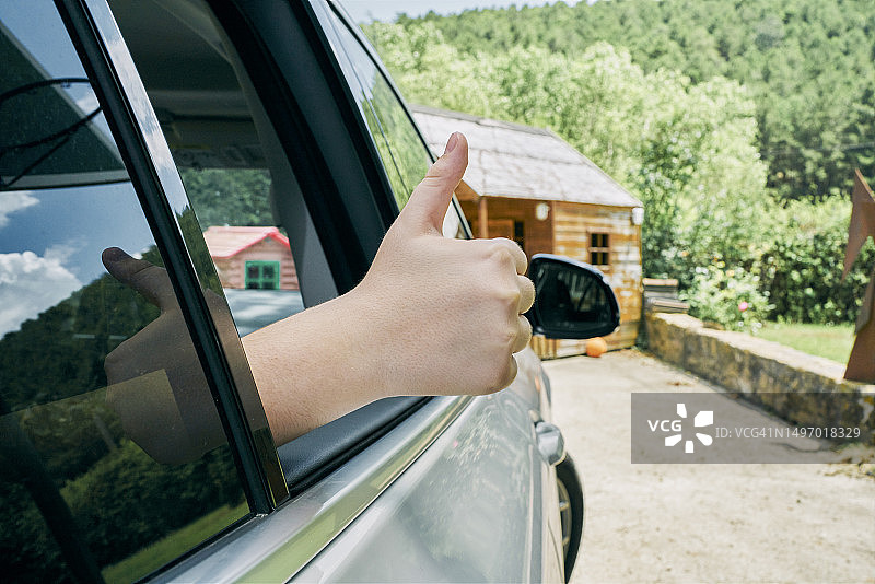 一只手在窗外一辆停着的汽车上竖起大拇指，只能看到汽车的一部分，在背景中可以看到前面的一所房子和绿地。图片素材