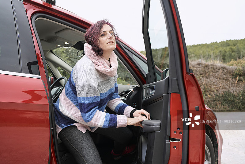 长途旅行后，一名穿着保暖衣服的女子从一辆红色轿车的副驾驶座位上下来休息。图片素材