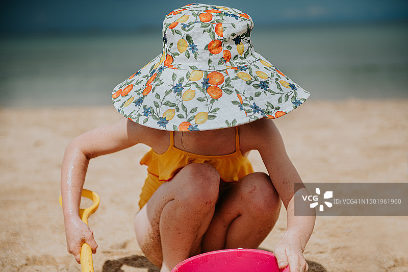 一个戴着宽边遮阳帽的小女孩蹲在沙滩上挖沙子图片素材
