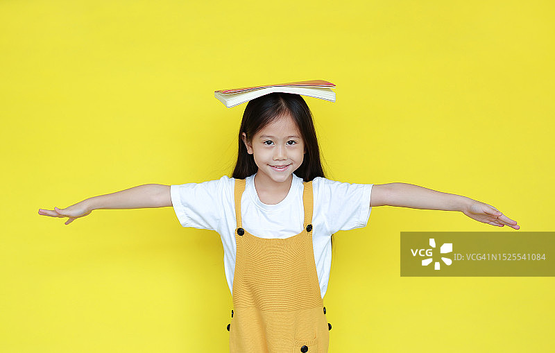 亚洲的小女孩，在黄色的背景上，头上顶着书。快乐的女学生的肖像，张开双臂，看着镜头。学生返校和作业概念，工作室短图片素材