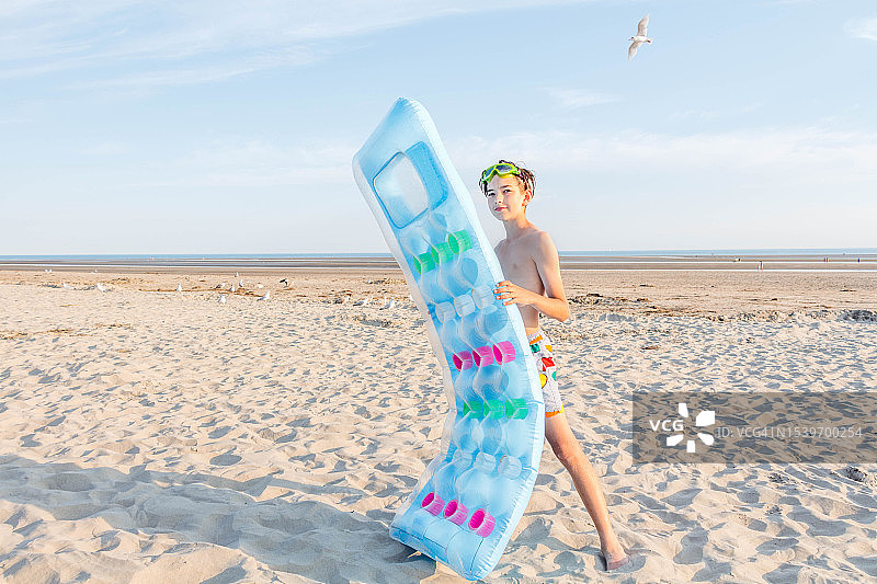一个男孩站在沙滩上，拿着充气床垫，面对着大海和蓝天图片素材