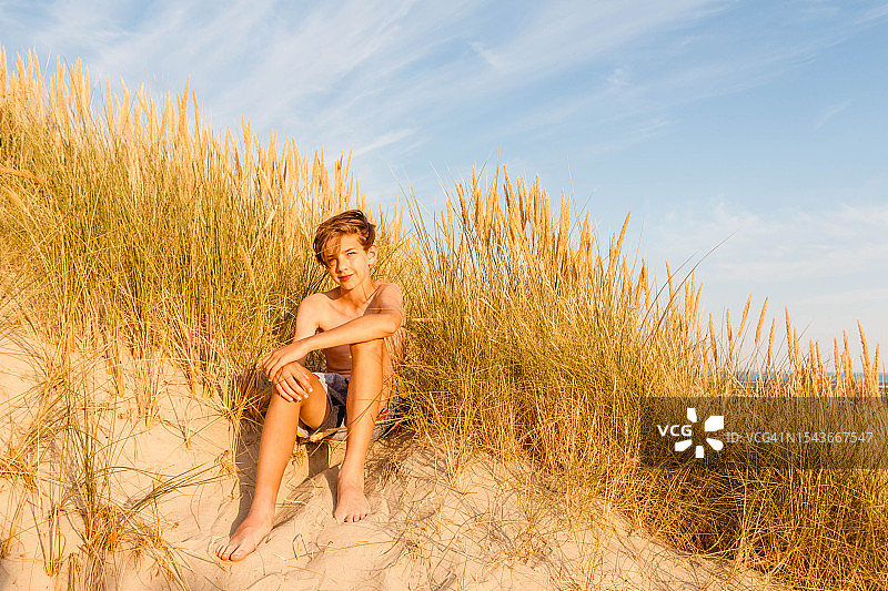 一个男孩在一个阳光明媚的日子里坐在沙丘上，背靠着一片红草坪图片素材