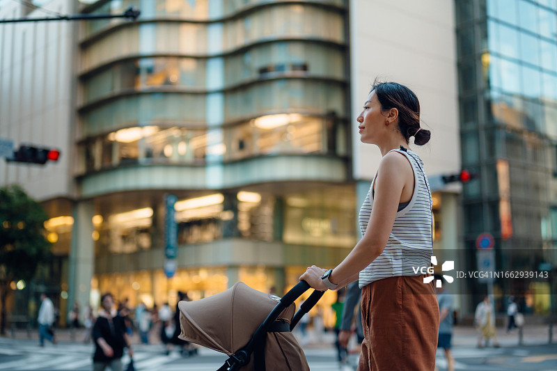 年轻的亚洲母亲推着婴儿车走在繁忙的城市街道上，对面是现代化的城市摩天大楼和购物中心。家庭生活方式。带宝宝逛街图片素材