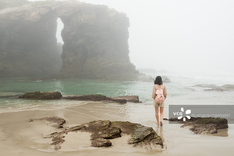 在一个雾蒙蒙的夏日里，西班牙加利西亚里巴迪奥，一名年轻女子穿着短裤，背着背包，穿过雄伟的大教堂海滩的岩层图片素材