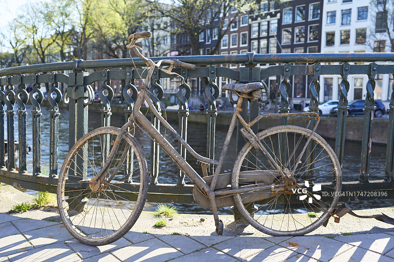 一辆生锈的旧自行车停在街道栏杆附近的人行道上，背景是阿姆斯特丹运河，侧视图图片素材