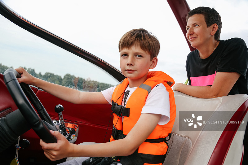 勇敢的8-9岁男孩穿着橙色救生衣训练驾驶快艇，迷人的妈妈为她的儿子感到骄傲图片素材