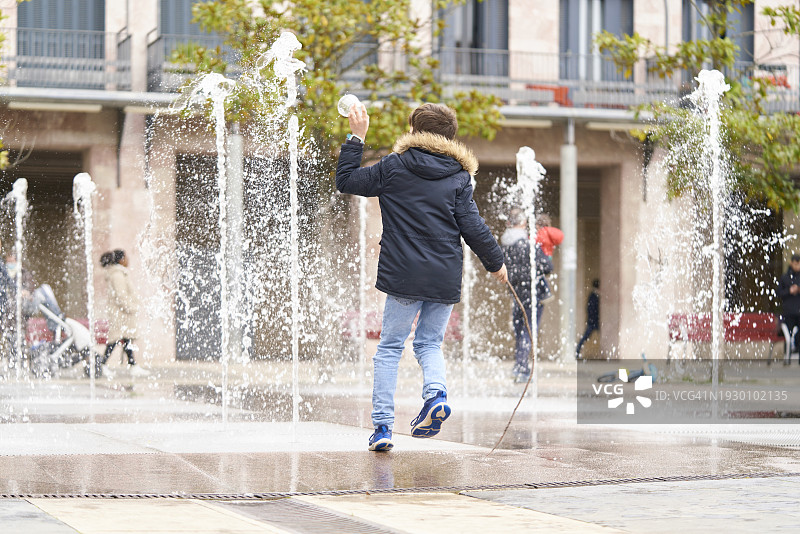 一个男孩的后视图，他在大楼外的街道喷泉中奔跑和玩耍图片素材