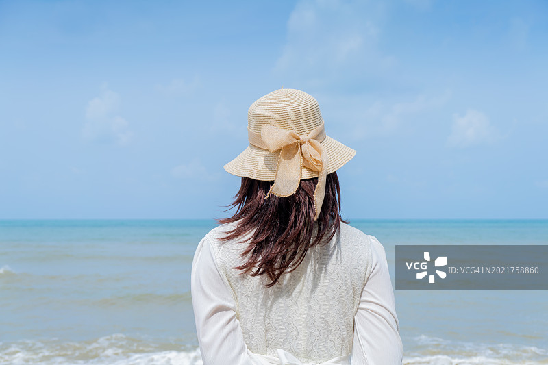 年轻女子穿着白裙子，戴着白帽子，站在沙滩上，双手伸向大海图片素材