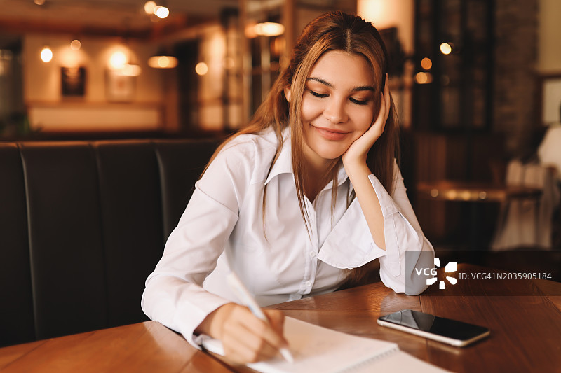 一个穿着白衬衫，微笑着恋爱的女孩坐在一个有着黑暗阁楼风格的咖啡馆的桌子旁写信。一名大学生用手机远程做作业和学习图片素材