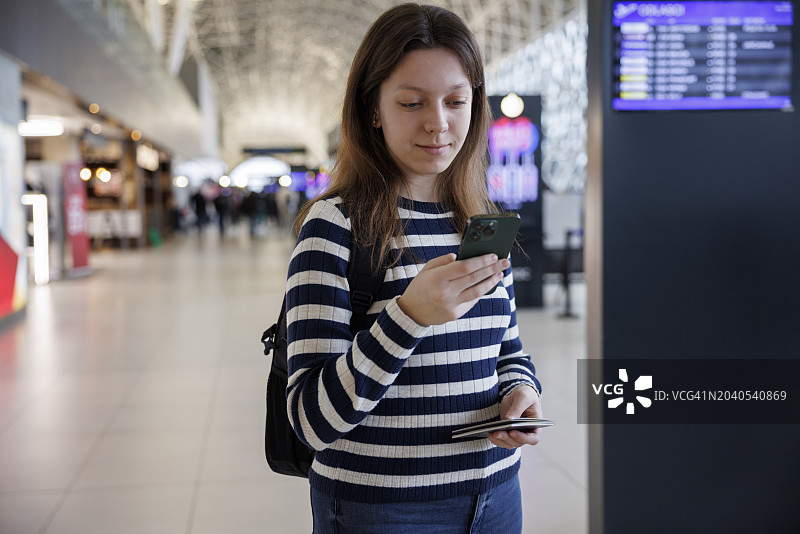 微笑的年轻女子背着背包在机场查看她的登机时间表图片素材