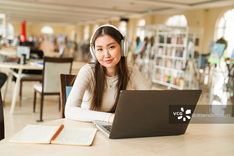 年轻的亚洲女人戴着耳机在图书馆用笔记本电脑学习图片素材