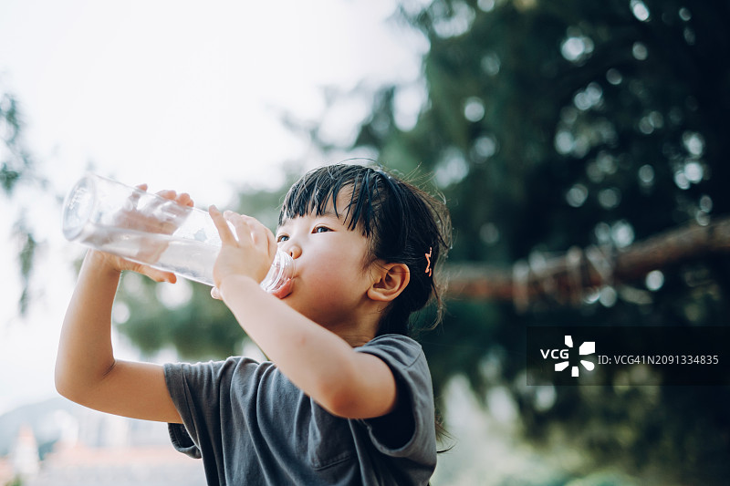 在一个阳光明媚的夏日，一个亚洲小女孩正在户外用可重复使用的塑料水瓶大口喝水。保持肌肤水润。健康生活方式理念图片素材