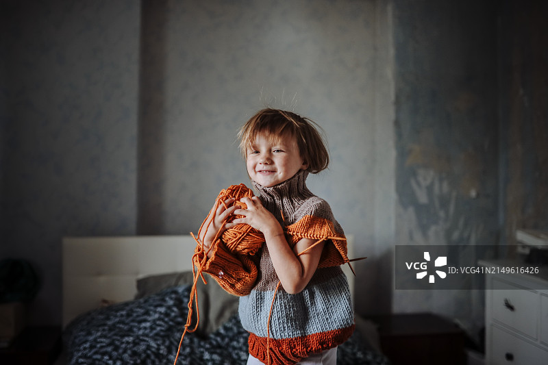 一个高兴的小女孩一边试穿一件未完成的毛衣，一边玩着纱线:创造性和愉快的活动，有意识的消费主义。图片素材