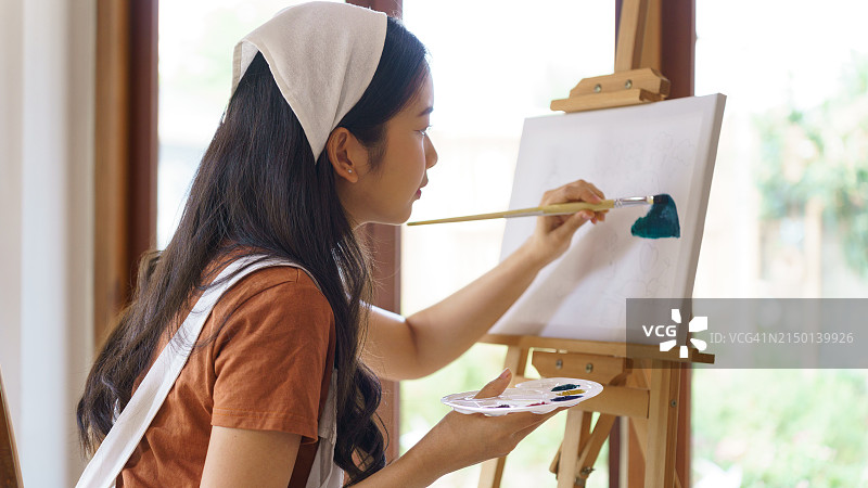 年轻的亚洲女艺术家手持调色板，用画笔在画架上绘制油画杰作，同时在工作室里以艺术爱好的生活方式创作艺术品图片素材