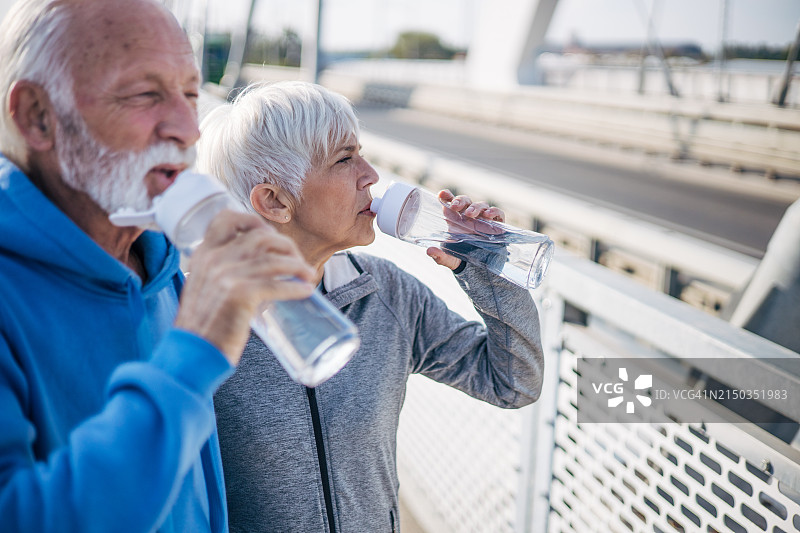 白天，一对老年夫妇在城市里运动后站在一起喝水图片素材