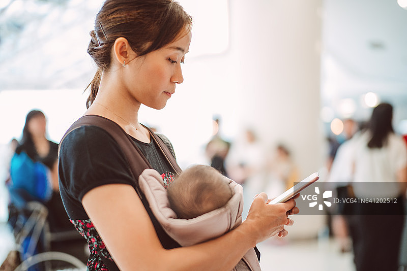 漂亮的年轻妈妈在购物中心用婴儿车抱着她的孩子，同时在用智能手机。图片素材