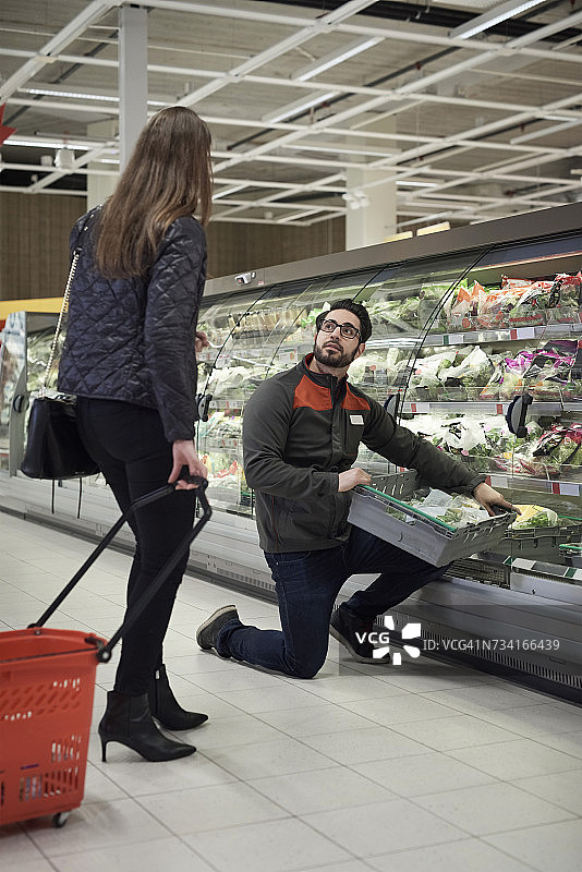 在超市里，一名男售货员跪在板条箱前，与一名提着篮子的女士交谈图片素材