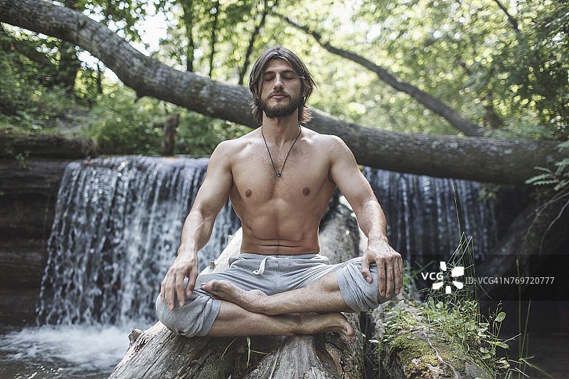 一名上身赤裸的男子，双腿交叉，坐在森林中瀑布旁的圆木上图片素材