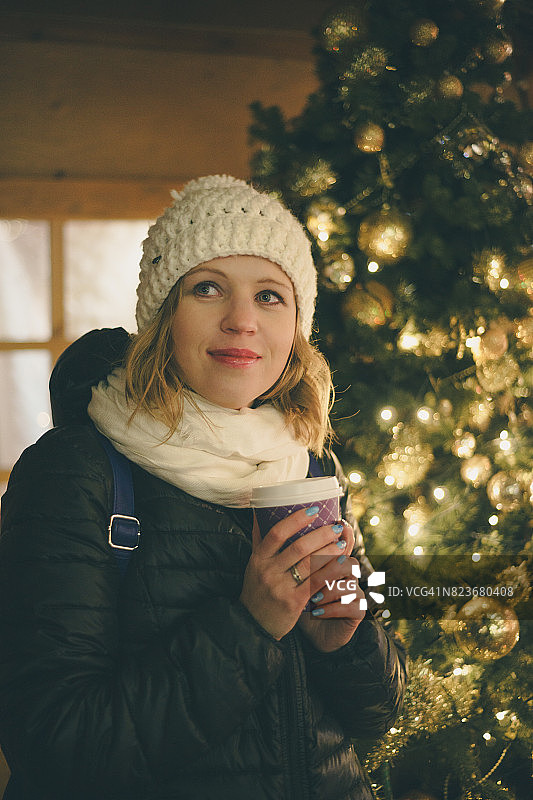 快乐的年轻女子在圣诞树旁的圣诞市场喝咖啡图片素材